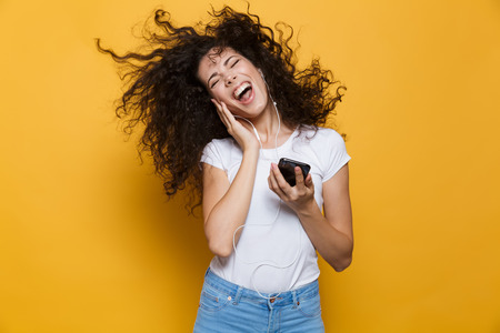 Image of happy woman 20s with curly shaking hair singing while holding smartphone and listening to...