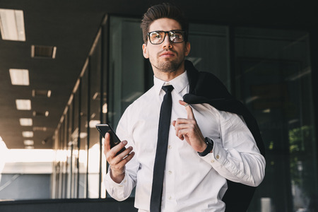 Image of businesslike man dressed in formal suit standing outside glass building with jacket over his shoulder and holding mobile phoneの写真素材
