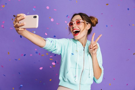Portrait of a happy young girl with bright makeup isolated over violet background, taking a selfie under confetti showerの写真素材