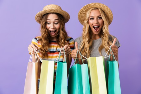 Two pretty happy young girls friends in summer hats standing isolated over violet background, looking inside shopping bagsの写真素材