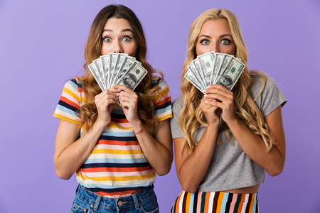 Two pretty young girls friends standing isolated over violet background, showing money banknotesの写真素材