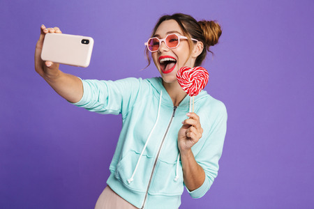 Photo of stylish young woman 20s with hair in buns holding lollipop and taking selfie isolated over...