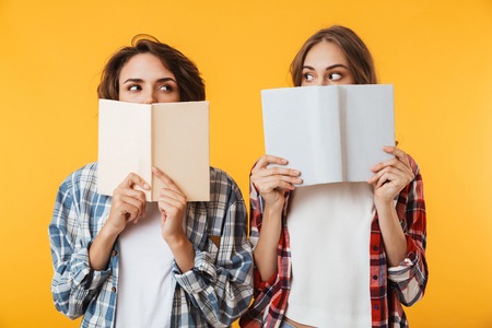 Photo of young women friends posing isolated over yellow background holding books covering faces.の写真素材