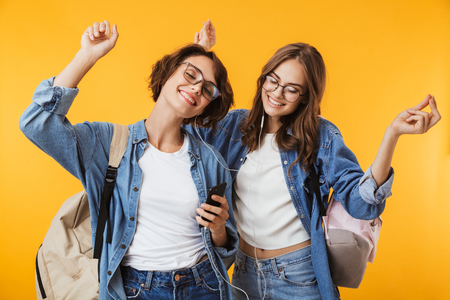 Image of cheerful happy women friends isolated over yellow wall background using mobile phone listening music with earphones.の写真素材