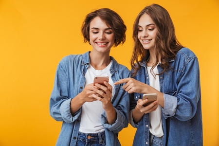 Photo of cheerful happy emotional young women friends posing isolated over yellow background using mobile phones.の写真素材
