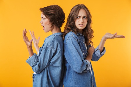 Photo of agressive displeased young women friends isolated over yellow background quarrel.