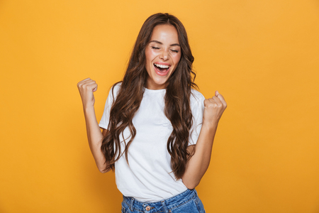 Portrait of a happy young girl with long brunette hair standing over yellow background, celebrating successの写真素材