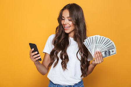 Portrait of a happy young girl with long brunette hair standing over yellow background, holding money banknotes, using mobile phoneの写真素材