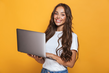 Portrait of pretty woman 20s with long hair smiling and holding gray laptop isolated over yellow backgroundの写真素材