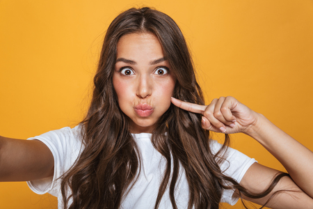 Portrait of excited woman 20s with long hair having fun while taking selfie photo isolated over yellow backgroundの写真素材