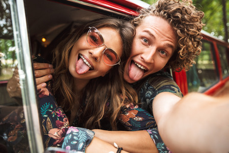 Photo of loving hippie couple smiling and showing peace sign while driving retro minivan in forest