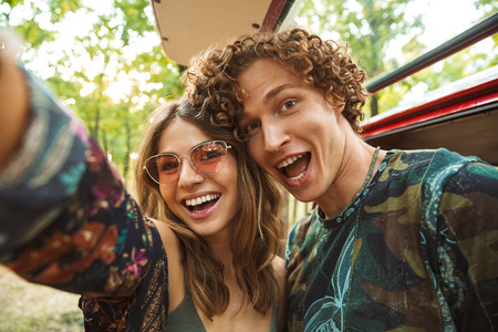 Photo of european hippie couple man and woman smiling and taking selfie in forest near retro minivanの写真素材