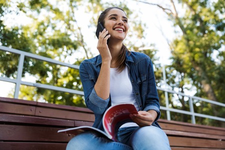 Image of amazing young beautiful woman sitting outdoors talking by mobile phone.の写真素材