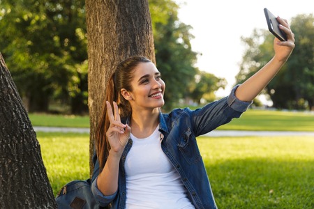 Image of amazing beautiful young woman student in the park take a selfie by mobile phone with peace gesture.の写真素材