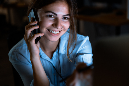 Cheerful young woman designer sitting in office working talking by mobile phone at night.