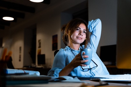 Image of cute young woman designer sitting in office working drinking coffee or tea at night.の写真素材