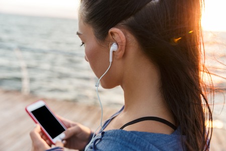 Image of beautiful young sports woman outdoors on the beach listening music with earphones using mobile phone.の写真素材