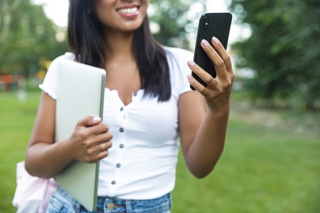 Cropped image of smiling asian student girl looking at mobile phone while holding laptop computer outdoorsの写真素材