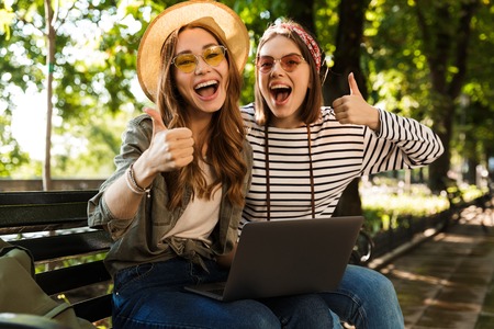 Photo of young emotional excited happy ladies friends outdoors sitting using laptop computer showing thumbs up gesture.の写真素材