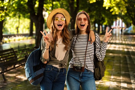 Image of excited beautiful young happy women friends walking outdoors with backpacks showing peace gesture.の写真素材