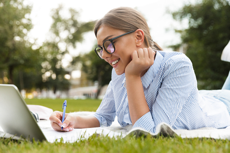 Cheerful young girl spending time at the park, studying, laying on a blanket with laptop computerの写真素材