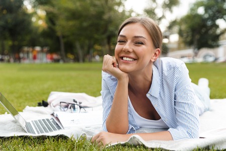 Cheerful young girl spending time at the park, studying, laying on a blanket with laptop computerの写真素材