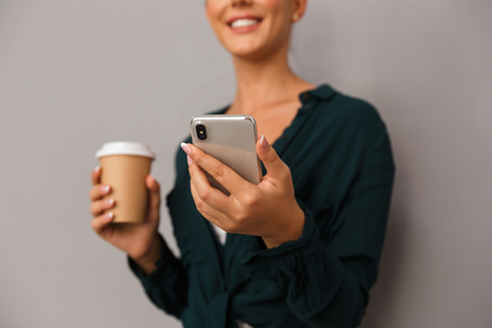 Cropped photo of excited beautiful business woman posing isolated over grey wall background drinking coffee talking by mobile phone.の写真素材