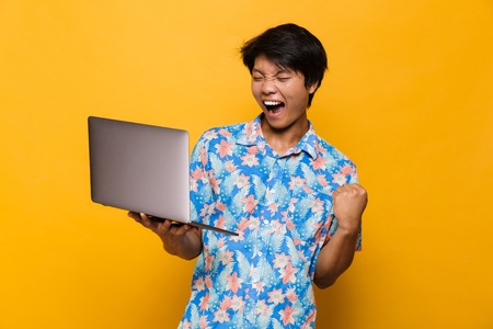 Image of a excited screaming young asian man standing isolated over yellow background using laptop computer make winner gesture.の写真素材