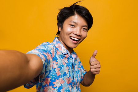 Image of happy young asian man posing isolated over yellow background take a selfie with thumbs up gesture.の写真素材