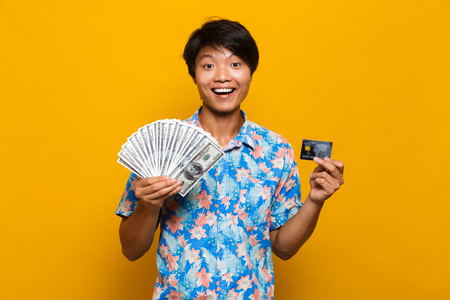 Image of happy young asian man standing isolated over yellow background holding money and credit card.の写真素材