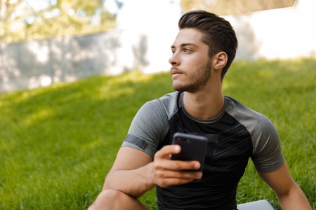 Image of young concentrated sports man outdoors in park looking aside using mobile phone.の写真素材