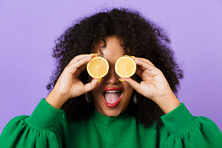 Photo of excited happy pretty african woman isolated over violet background holding lemons covering eyes.の写真素材