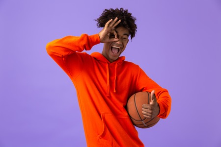 Portrait of a cheerful young african man holding a basketball isolated over violet background, showing okの写真素材