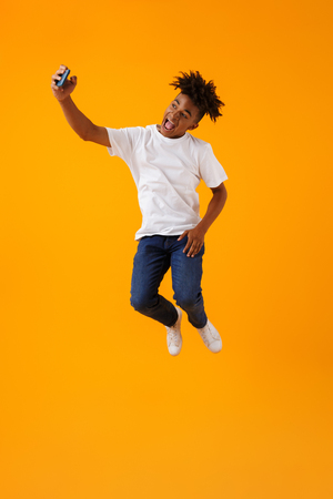 Image of happy young african man jumping isolated over yellow background take a selfie by phone.の写真素材