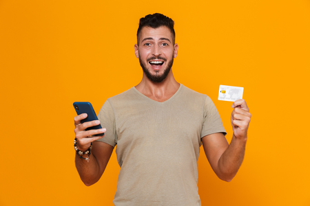 Portrait of an excited young bearded man in t-shirt isolated over orange background, holding mobile phone, showing plastic credit cardの写真素材