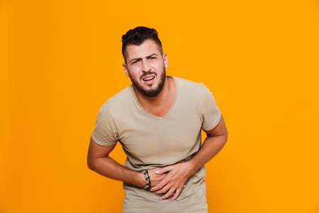 Portrait of a young casual man standing isolated over orange background, suffering from a strong stomach acheの写真素材