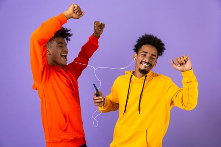 Two joyful african man friends dressed in colorful hoodies standing isolated over violet background, listening to music with earphones and mobile phone, dancingの写真素材