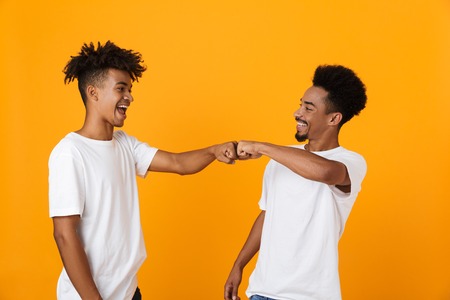 Two happy male african friends in t-shirts standing isolated over yellow background, giving fist bumpの写真素材
