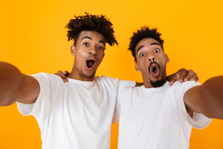 Two joyful male african friends in t-shirts standing isolated over yellow background, taking a selfieの写真素材