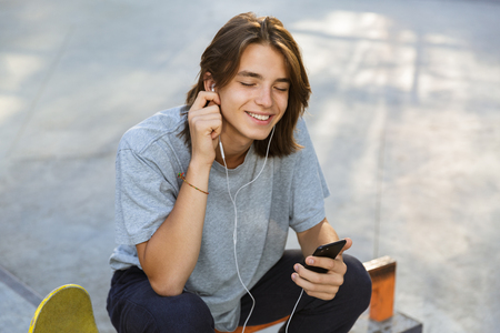 Joyful young guy spending time at the skate park, listening to music with earphones