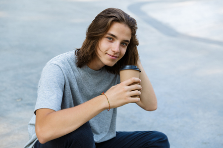 Happy young teenge boy spending time at the skate park, sitting on a skateboard, holding takeaway coffeeの写真素材