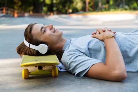 Smiling young guy spending time at the skate park, listening to music with headphones, laying on...