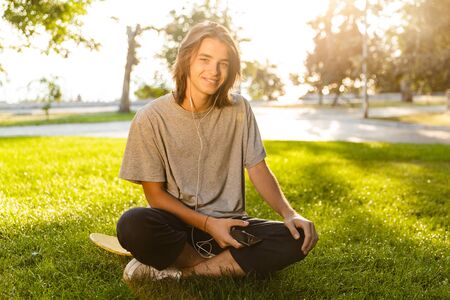 Image of handsome young guy sitting outdoors in the park on grass looking camera listening music with earphones.の写真素材