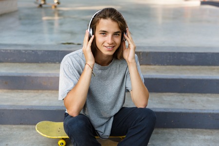 Smiling young teenage boy spending time at the skate park, listening to music with headphones, sitting on skateboardの写真素材