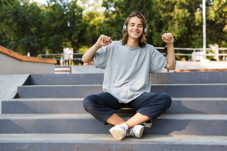 Smiling young teenage boy spending time at the skate park, listening to music with headphones, sitting on skateboard, dancingの写真素材