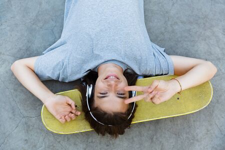 Top view of a young guy spending time at the skatepark, listening to music with headphones, laying on skateboard, showing peaceの写真素材