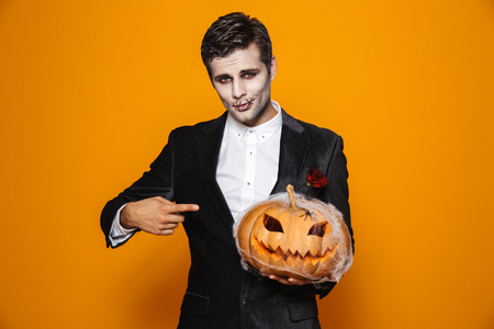 Photo of terrifying dead man on halloween wearing classical suit and creepy makeup holding carved pumpkin isolated over yellow backgroundの写真素材