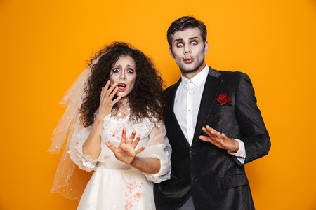 Photo of terrifying zombie couple bridegroom and bride wearing wedding outfit and halloween makeup scaring you isolated over yellow background