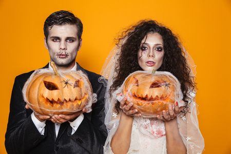 Photo of scary zombie couple bridegroom and bride wearing outfit and halloween makeup holding carved pumpkin isolated over yellow backgroundの写真素材