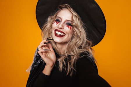 Portrait of blonde scary woman witch in black hat looking camera and smiling isolated over orangeの写真素材